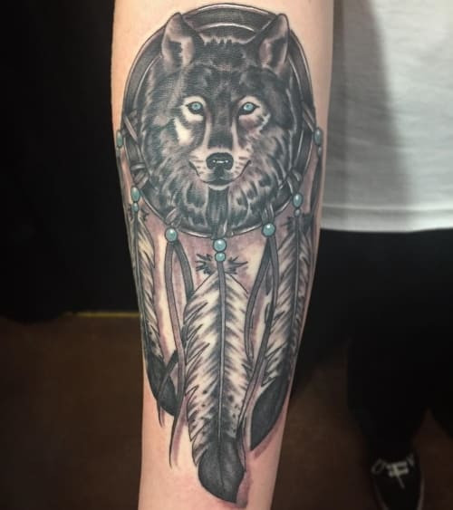 A wolf dreamcatcher tattoo on a man's forearm, merging a detailed wolf image with an intricate dreamcatcher design, symbolizing protection from negativity and promotion of positive energy.