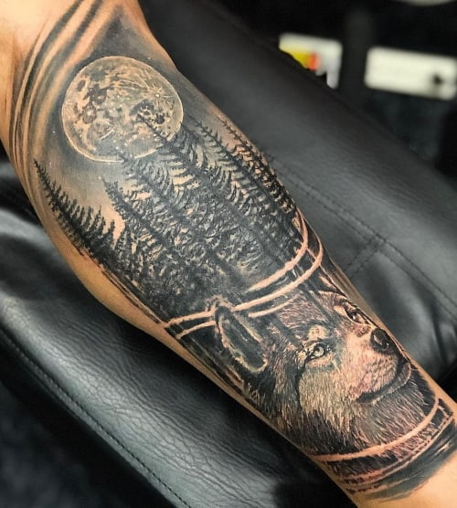 A wolf in the woods tattoo on a man's thigh, featuring a wolf within a detailed forest scene, symbolizing connection to nature, peace, rejuvenation, and inner healing.