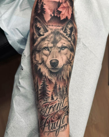 Detailed wolf head tattoo on a man's outer forearm, featuring realistic fur texture, piercing eyes, and shading in black and grey.