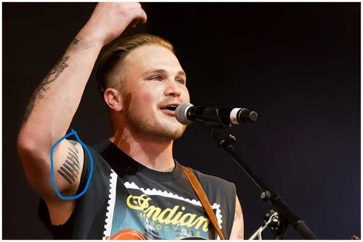 Zach Bryan performing during the Quittin Time tour, showing his guitar strings tattoo.