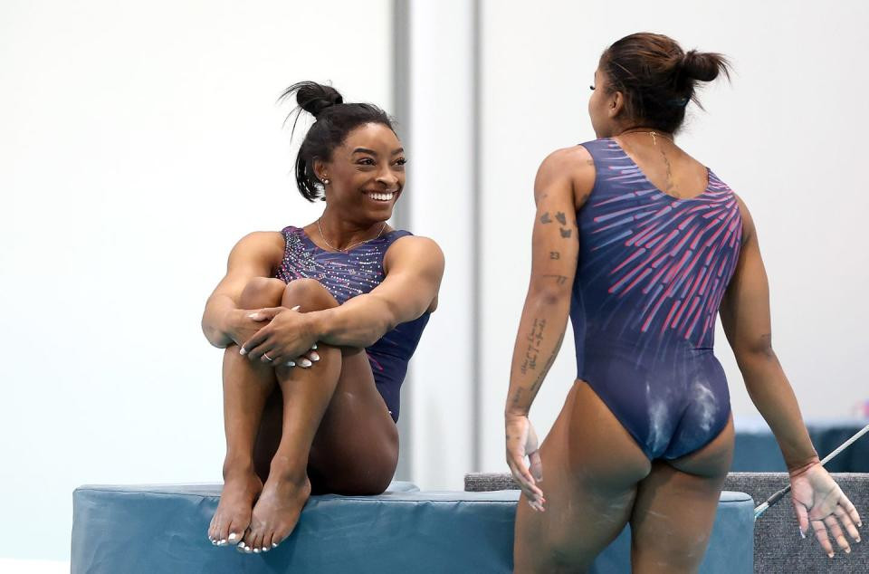 simone biles smiles at jordan chiles as she sits on a gymnastics mat
