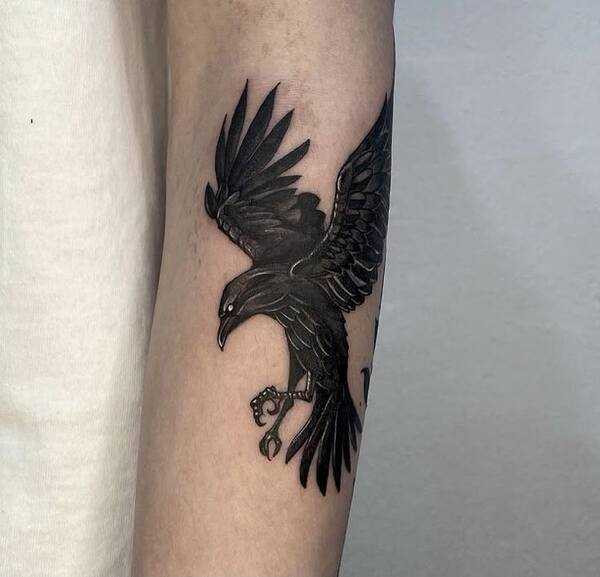 A crow tattoo design placed on the forearm, featuring intricate linework and feather details