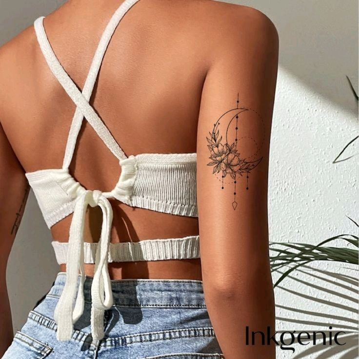 Woman with moon and flower tattoo on upper arm wearing a white strappy top and jeans, standing against a white wall.