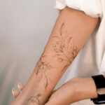 Close-up of a forearm with a detailed floral tattoo, featuring delicate flowers and leaves, person wearing a smart watch.