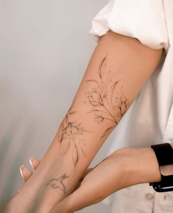 Close-up of a forearm with a detailed floral tattoo, featuring delicate flowers and leaves, person wearing a smart watch.