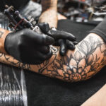 girl with dreadlocks in a tattoo parlor. The master creates a picture on the body of a young beautiful girl. Close-up of hands and tattoo machine
