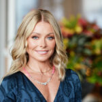 Kelly Ripa smiling while wearing a black dress with a small tattoo visible on her inner arm