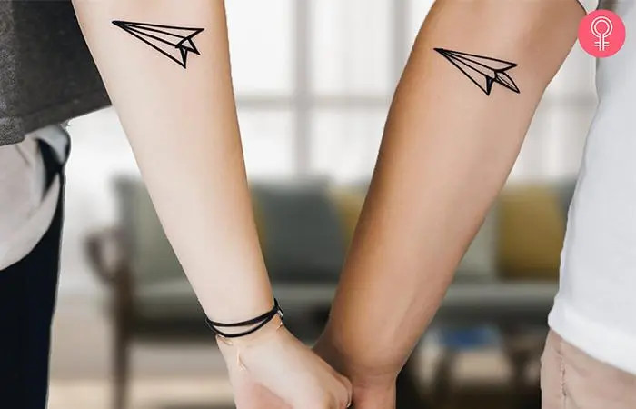 Two hands display matching paper airplane tattoos, symbolizing a shared journey.