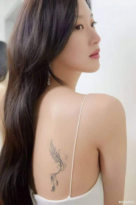 Woman with long dark hair and winged tattoo on back, wearing a white strap dress, glancing over her shoulder.