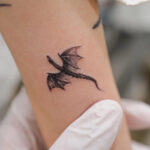 A person's wrist adorned with a minimalist black ink tiny dragon tattoo, showcasing the elegance of simplicity in design.