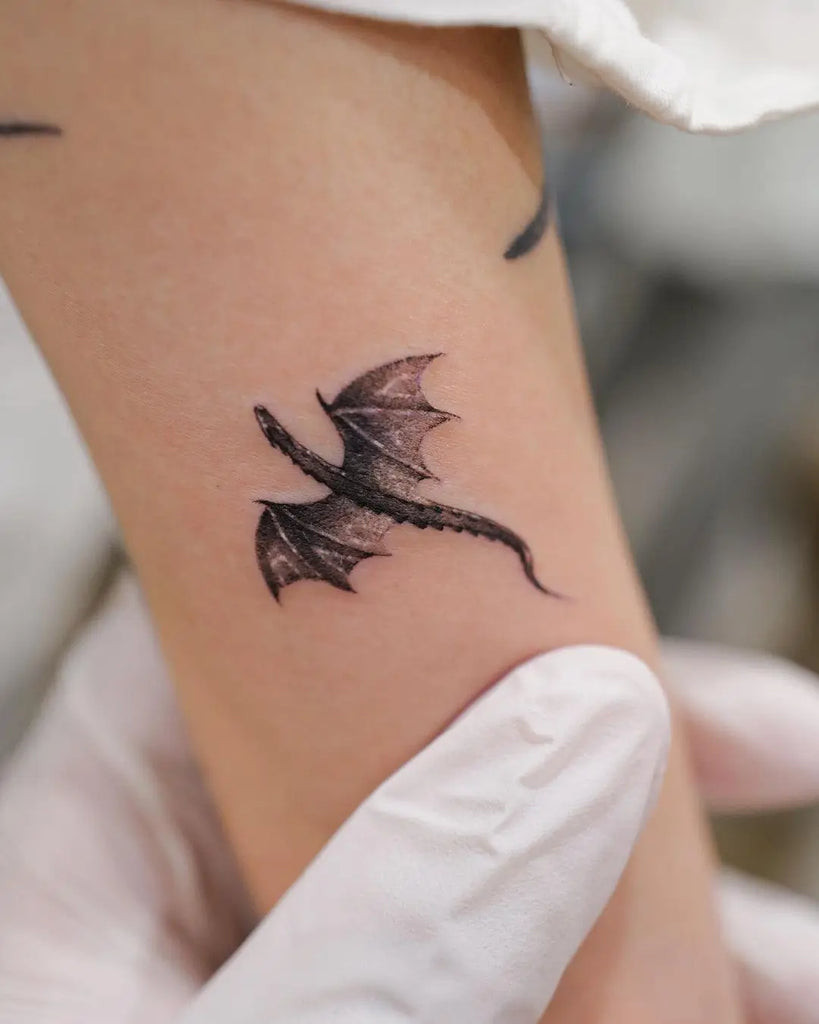 A person's wrist adorned with a minimalist black ink tiny dragon tattoo, showcasing the elegance of simplicity in design.