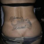 Woman's lower back tattoo with a huge throbbing cock encompassed by the words Woman's best friend