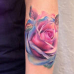 Vibrant watercolor rose tattoo on an arm, showcasing detailed petals in shades of pink, purple, and blue.