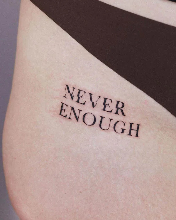 Quote tattoo Never Enough on the thigh, by @baekryeon_tattoo, symbolizing ambition and self-drive