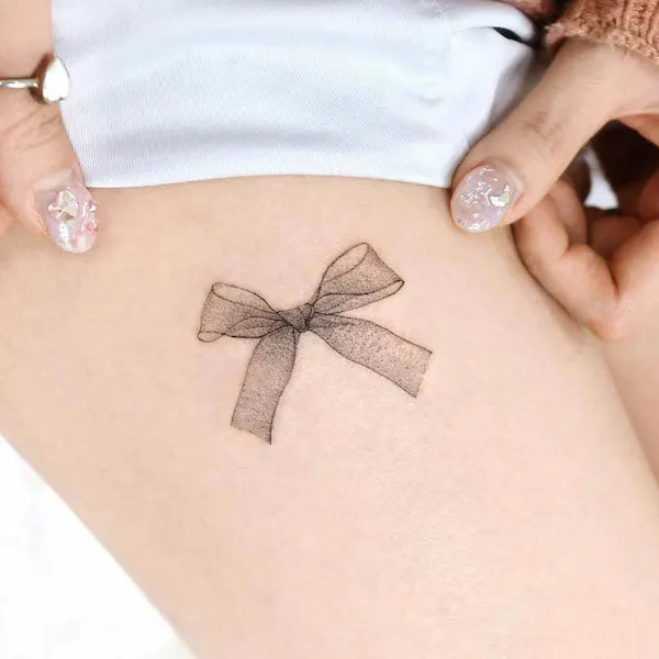 Delicate bow tattoo on the thigh, by @palette.tt_, representing femininity and charm