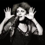 Stevie Nicks sings into a microphone and holds her hands near her face.